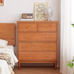 Front view of Manna cherry wood 5-drawer chest of drawers styled beside a bed in a bright, cozy bedroom.