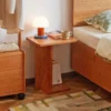 Manna cherry wood C-shaped side table placed beside a bed as a nightstand with a small lamp and books.