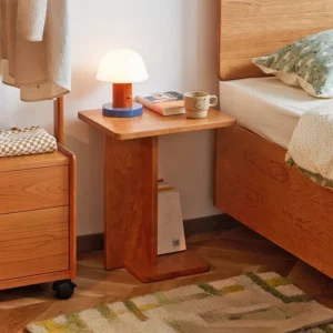 Manna cherry wood C-shaped side table placed beside a bed as a nightstand with a small lamp and books.