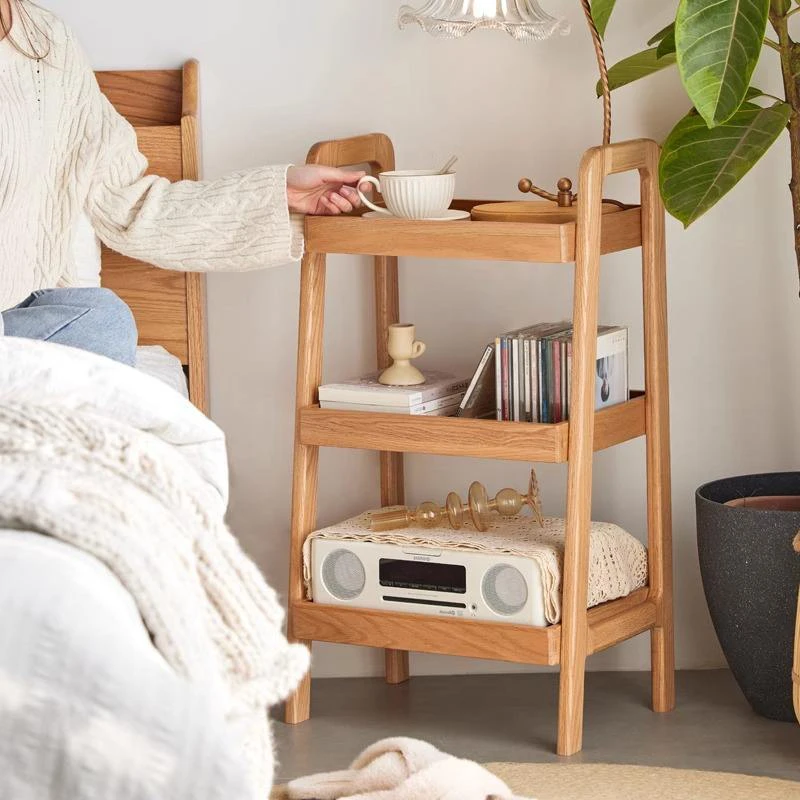 Manna oak 3-tier ladder nightstand with tray shelves styled beside a bed holding a teacup, books, and CD player.