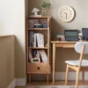 Manna oak narrow glass bookcase placed next to a wooden writing desk in a home office nook holding books and a camera.