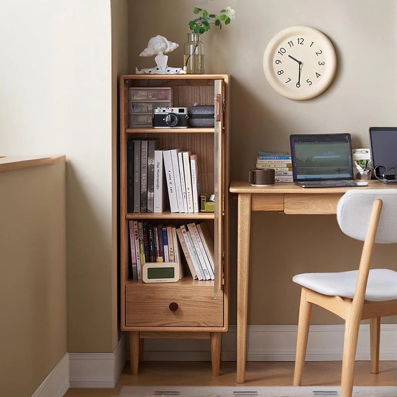 Manna oak narrow glass bookcase placed next to a wooden writing desk in a home office nook holding books and a camera.