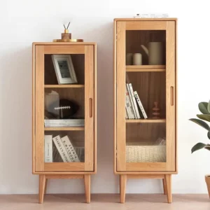 Front view comparison of Manna oak narrow glass display cabinets in 1000mm and 1150mm heights holding books, photo frame, and tea set.