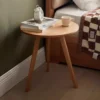 Manna oak round side table with 3 tapered legs placed beside a bed in a cozy bedroom with books and a white alarm clock.