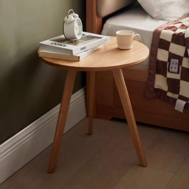 Manna oak round side table with 3 tapered legs placed beside a bed in a cozy bedroom with books and a white alarm clock.