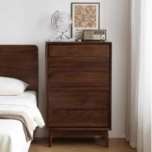 Front view of Manna black walnut 5-drawer tall chest placed next to a bed in a warm bedroom.