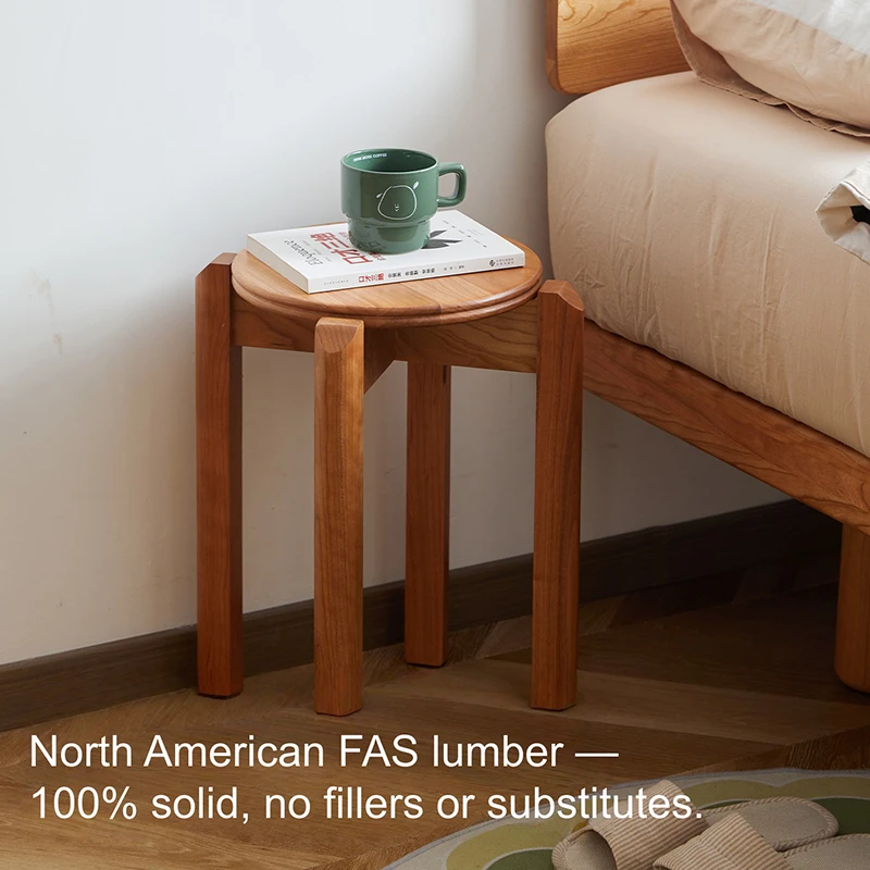 manna-cherry-stackable-stool-lifestyle Round solid cherry wood stool with a green ceramic mug and book on top beside a natural wood bed in a bedroom.