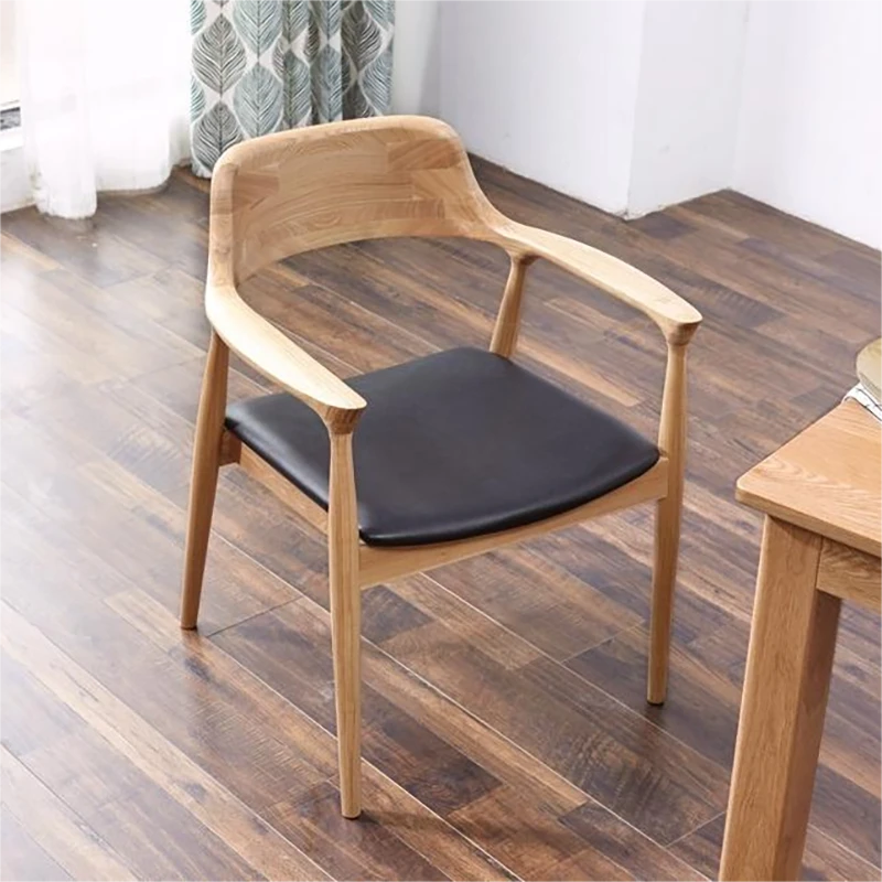 Solid oak armchair with a black leather seat cushion placed on dark wood flooring in a bright interior setting.