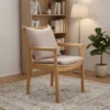 manna-oak-armchair-spindle-back-living-room Lifestyle view of solid oak dining armchair with spindle back and beige cushion in a living room beside a bookshelf and floor lamp.