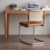 Lifestyle view of an oak cane back cantilever chair with a tan leather seat at a wooden desk with a laptop and clock.