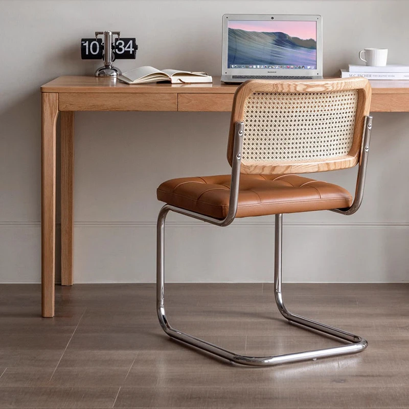Lifestyle view of an oak cane back cantilever chair with a tan leather seat at a wooden desk with a laptop and clock.
