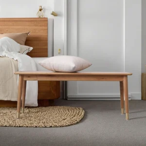 Oak bench at the foot of a bed with a pink cushion and brass capped tapered legs on a grey carpet.
