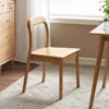 Back view of an oak dining chair with a curved open backrest and solid wood seat next to a light oak chest of drawers.