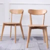manna-oak-dining-chair-curved-back-pair Two solid oak dining chairs with curved wide backrests and solid wood seats photographed against a white paneled wall.