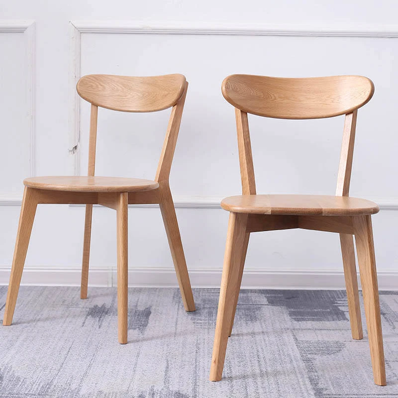 manna-oak-dining-chair-curved-back-pair Two solid oak dining chairs with curved wide backrests and solid wood seats photographed against a white paneled wall.