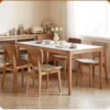 Lifestyle view of six oak dining chairs with upholstered backs around a white stone dining table in a bright dining room.