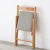 Back view of a light oak folding chair folded flat against a white wall showing the grey upholstered cushion.