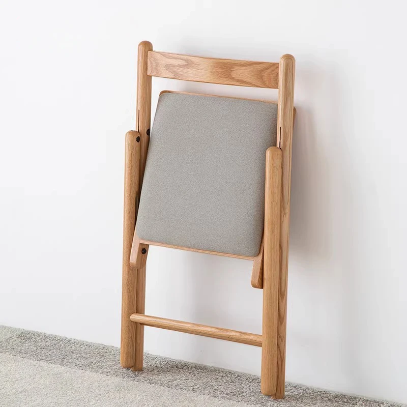 Back view of a light oak folding chair folded flat against a white wall showing the grey upholstered cushion.