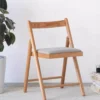 Front view of a light oak folding dining chair with a solid wood backrest and grey fabric seat cushion on a carpet.
