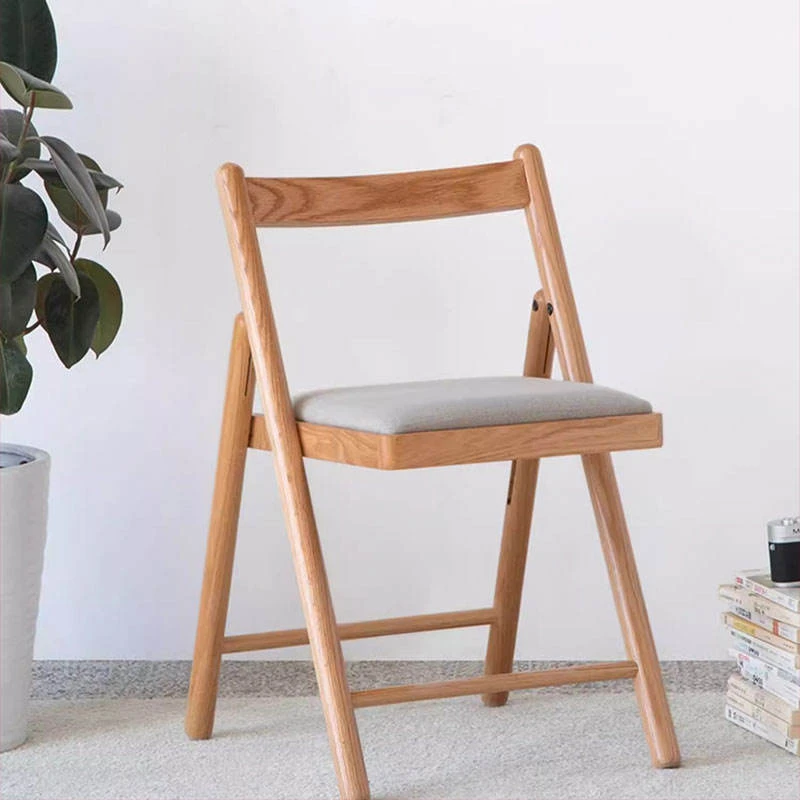 Front view of a light oak folding dining chair with a solid wood backrest and grey fabric seat cushion on a carpet.