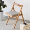Light oak folding dining chair with a grey fabric upholstered seat cushion on a rug beside a potted plant and books.