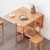 Lifestyle scene of a light oak folding dining table and stool set with one leaf raised beside a white wall.