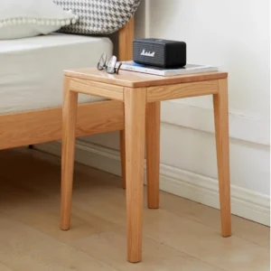 Solid oak square stool used as a bedside table holding a speaker, glasses and book beside a wooden bed.