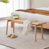 manna-oak-rattan-dining-bench-bedroom Oak rattan cane bench placed at the foot of a bed in a bright bedroom with a grey rug and linen bedding.