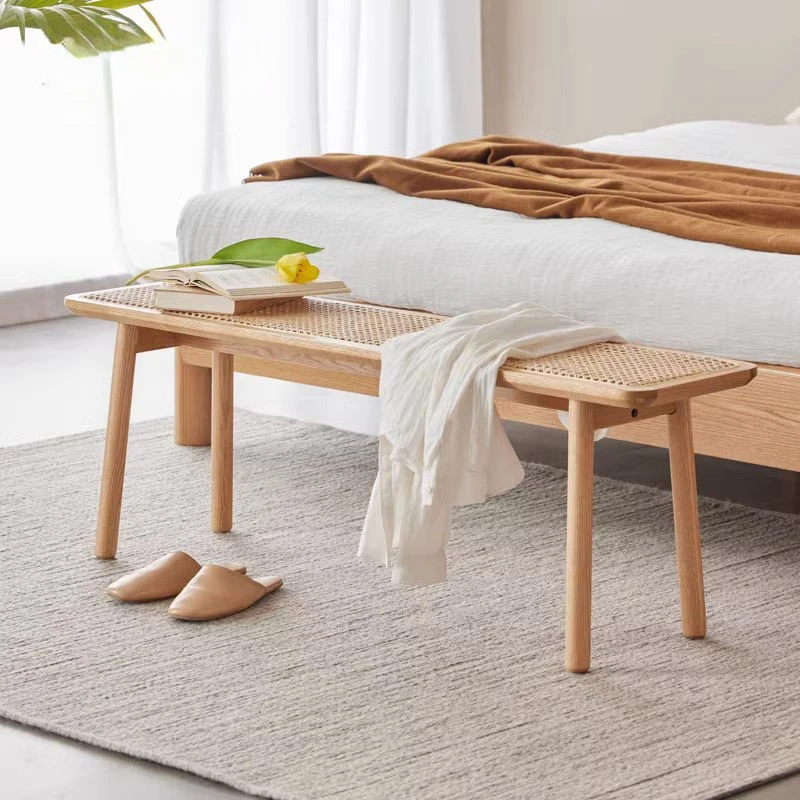 manna-oak-rattan-dining-bench-bedroom Oak rattan cane bench placed at the foot of a bed in a bright bedroom with a grey rug and linen bedding.