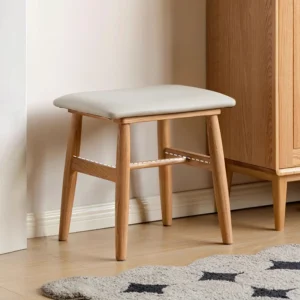 Oak square stool with light grey upholstered cushion seat standing beside a tall oak wardrobe on pale flooring.