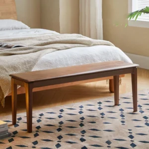 Walnut dining bench used at the foot of a bed on a patterned rug with layered linen bedding in a warm bedroom.