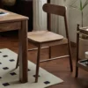 Side view of a solid walnut dining chair with a curved backrest and wooden seat beside a walnut table on herringbone flooring.