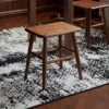 manna-walnut-square-stool-front-view Walnut square stool with a rectangular seat and four wooden screw plugs shown beneath a dining table on a rug.