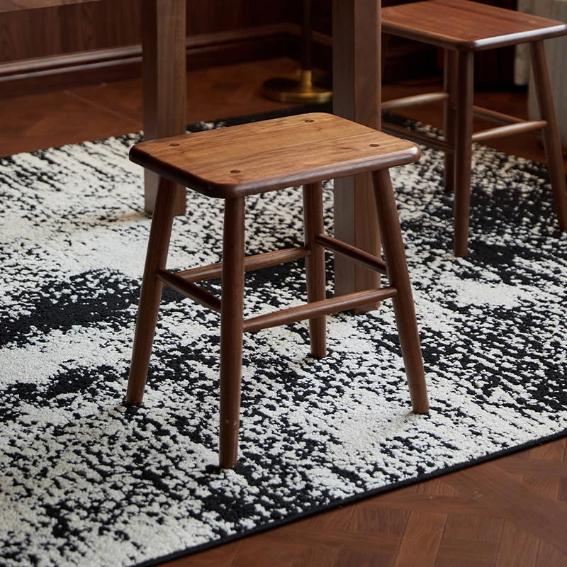 manna-walnut-square-stool-front-view Walnut square stool with a rectangular seat and four wooden screw plugs shown beneath a dining table on a rug.