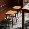 manna-walnut-square-stool-pair-lifestyle High angle view of two solid walnut square stools with rounded seats and angled legs on a black and white rug.