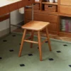 manna-walnut-square-stool-study-lifestyle Dark walnut square stool with splayed legs placed in a study beside wooden cabinetry on a green geometric rug.